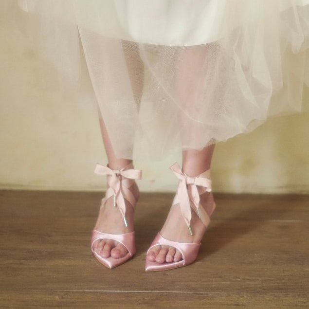 Pink satin shoes with ribbon ties worn by a person in a white dress on a wooden floor.