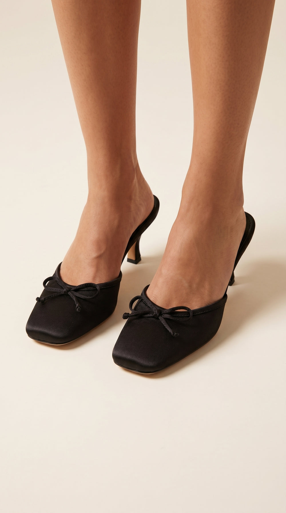 Black heeled shoes with bow details worn by a person on a beige background