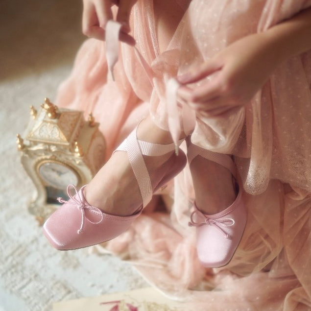 Person wearing pink ballet shoes with ribbons, sitting on a textured surface.