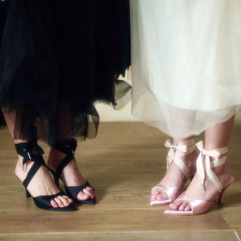 Two pairs of feet wearing black and pink high-heeled shoes on a wooden floor.